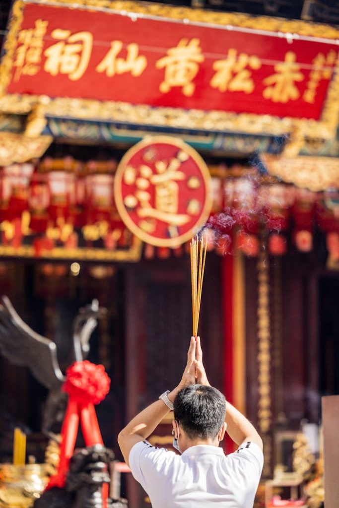 man-in-chinese-street-standing-and-holding-thin-candles-in-raised-arms-9839895 Rear view of a man holding incense in a religious ceremony at a Chinese temple with red and gold decorations.